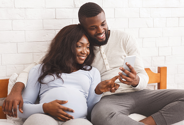 Cheerful pregnant black woman and her husband using phone, laying on bed, having good time together