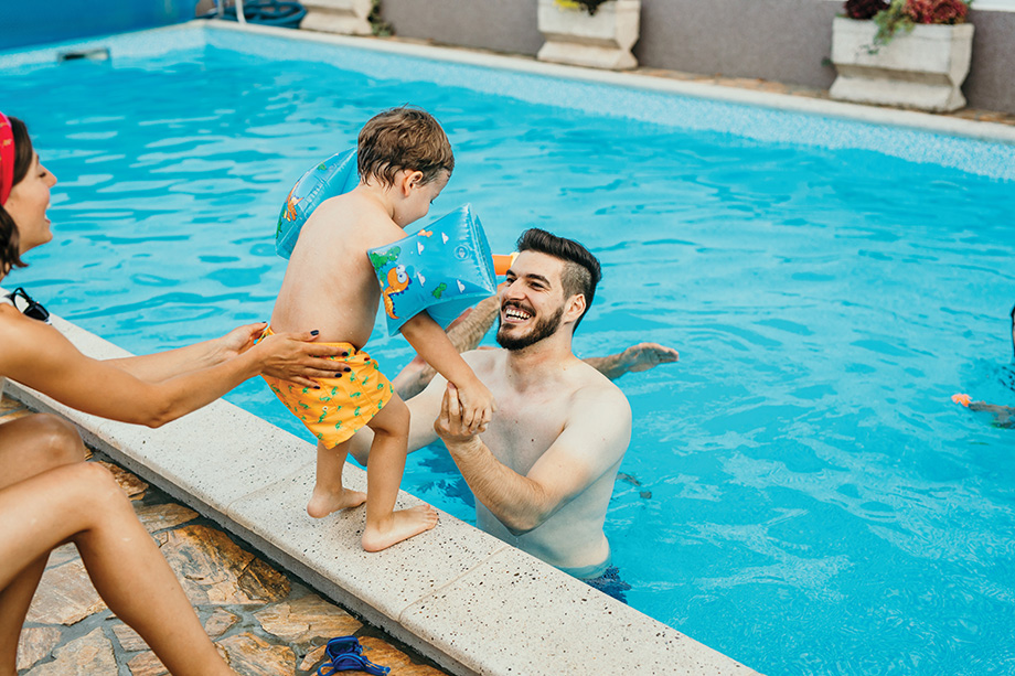 Fun, spontaneous young adult parents, playing at their summer house, at the pool. A father is inside the pool with their two years old sun, while cute looking mom, a brunet, is passing the son to her husband. All smiling, enjoying, and having fun