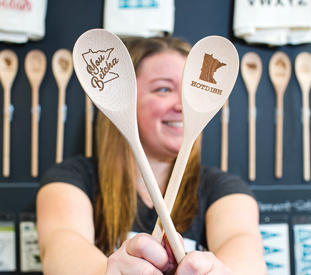 A woman holds two wooden serving spoons with a Minnesota theme from The Vintage Studio