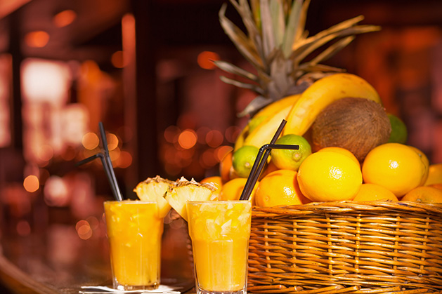 Two glasses of a summertime alcoholic punch rest on a bar in front of a basket of fresh fruit.