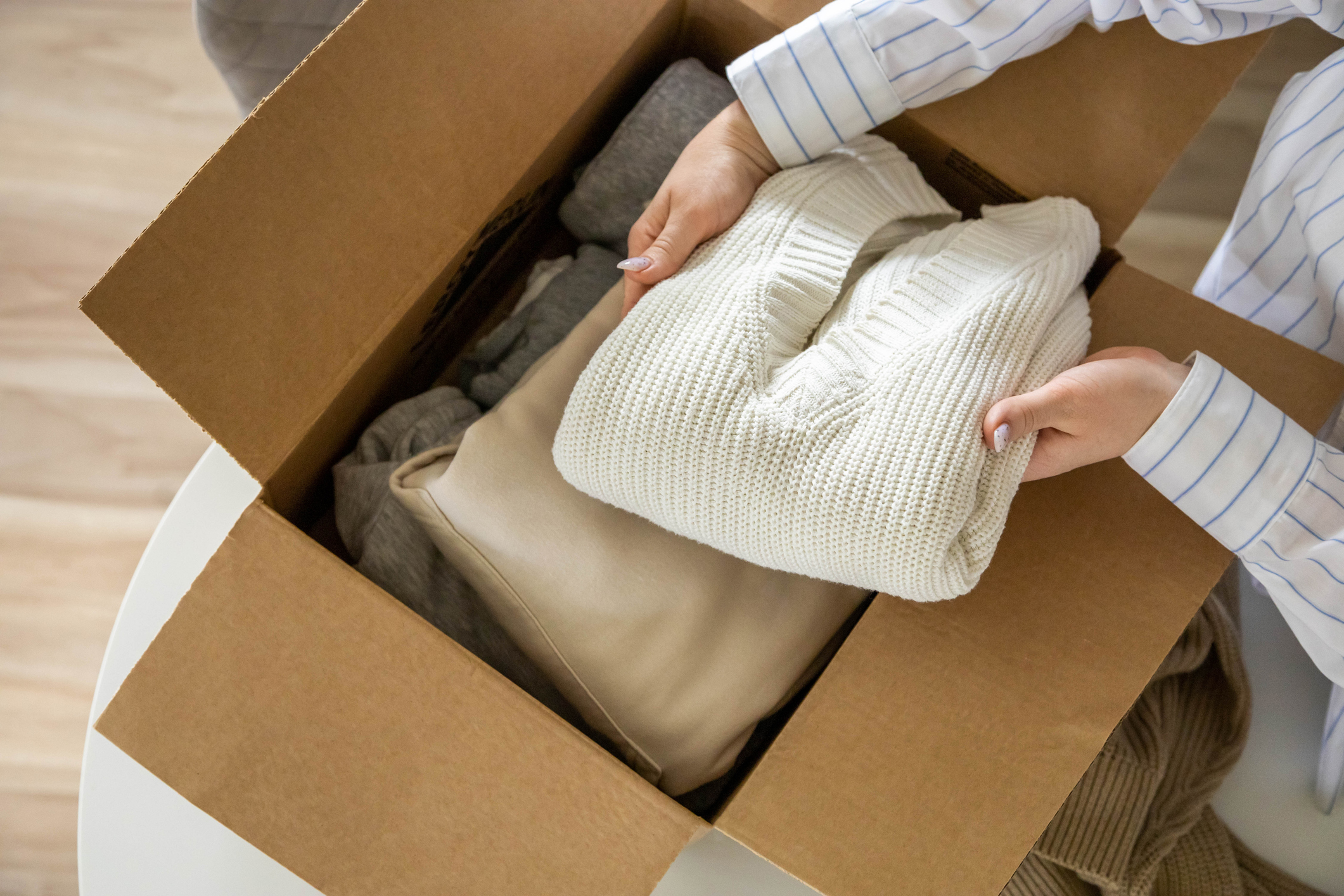 Woman hands putting warm neutral sweater into cardboard box seasonal storage method and donation closeup. Female arms packing taking knitted clothes apparel charity online shopping order tidying up