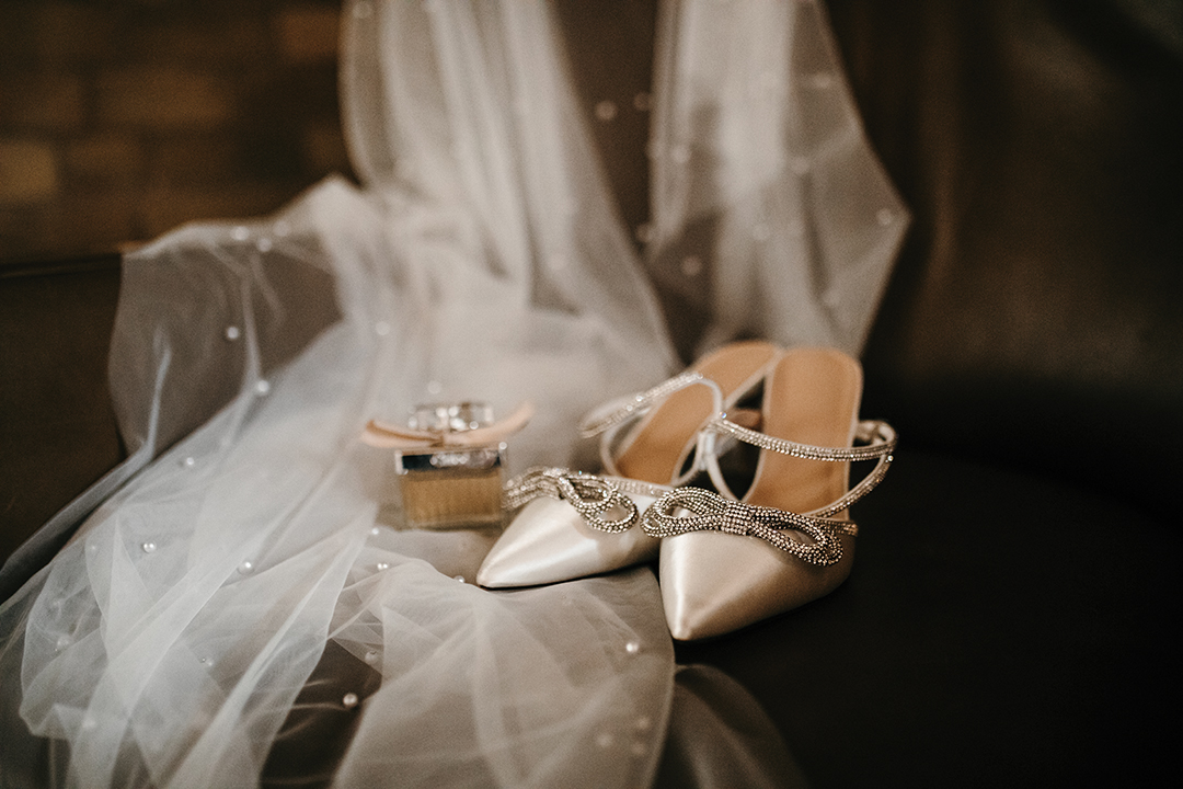 The bride’s lustrous satin gown from Vow’d Weddings was altered and customized by Heather Baldridge from Custom Sewing by Heather, who added satin buttons—flown in from Serbia—down the back of the gown. The look was paired with a chapel-length, angel-cut pearl veil from Tailored Tulle, pearl-drop earrings and white, satin heels, featuring dazzling bows. In the evening, the veil was exchanged for pearl gloves, also from Tailored Tulle.